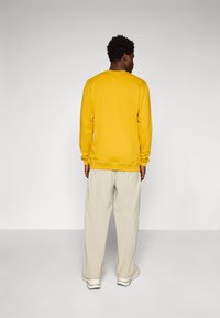 Sweatshirt jaune avec des poignets et un ourlet côtelés, associé à un pantalon large beige clair et des baskets blanches, vu de dos.