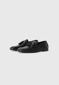 Une paire de mocassins en cuir noir avec des franges et des talons bas sur un fond blanc, légèrement inclinés vers la gauche.