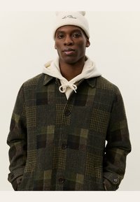 Cappotto di lana a quadretti verdi con colletto, bottoni neri e tasche laterali, indossato sopra un hoodie grigio. Un berretto di maglia bianca completa il look.