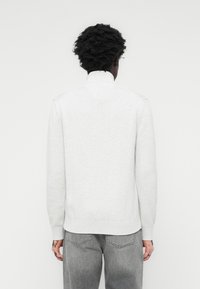 Lys grå strikkegenser med høy krage, lange ermer og ribbede mansjetter og kant. Stoffet ser mykt og teksturert ut.