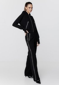 Czarny komplet z huedą i spodniami, z zamykanymi kieszeniami, dopasowanym krojem i kontrastującymi akcentami zamków. Materiał wydaje się miękki i elastyczny.