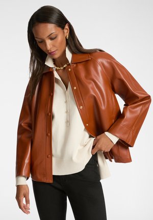 Giacca in finta pelle marrone con chiusura a bottoni, vestibilità oversize e ampio colletto. Abbinata a un maglione crema e pantaloni neri. Dettagli in metallo dorato.
