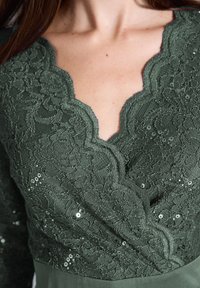 Robe en dentelle avec un décolleté en V profond aux bords festonnés, de couleur vert foncé, ornée de motifs floraux complexes et de paillettes dispersées pour la texture.