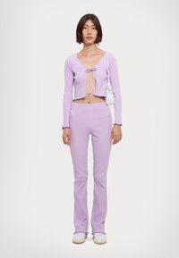 Lilac ribbed crop top with a tie front detail, paired with matching ribbed flared pants. Model holds a textured white handbag.