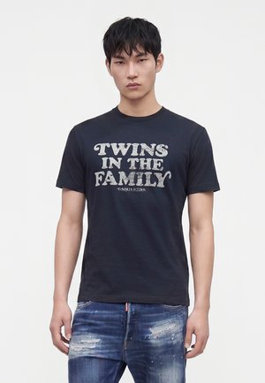 Jeune homme aux cheveux noirs courts porte un T-shirt noir avec l'inscription "Twins in the Family", associé à un jean bleu déchiré sur un fond uni.