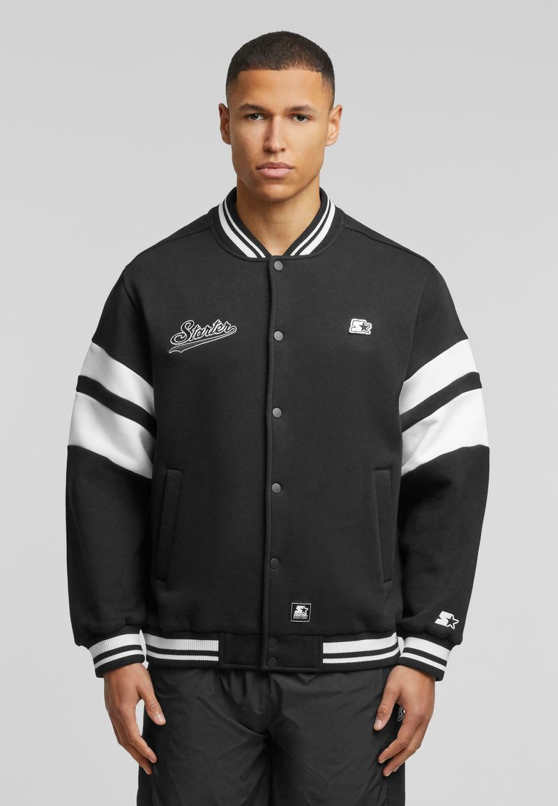 Black varsity jacket with white striped sleeves, snap buttons, embroidered logo, and ribbed cuffs and hem. Made from a soft fabric.