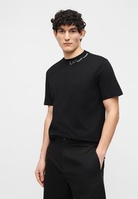 Νεαρός με σγουρά μαλλιά που φοράει μαύρο πουκάμισο με κοντά μανίκια και το κείμενο "EA7 Emporio Armani" κοντά στον γιακά, στέκεται με το ένα χέρι στην τσέπη.