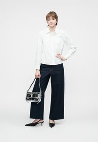 Camicia bianca a maniche lunghe con bottoni, jeans ampi blu scuro a vita alta, scarpe nere a punta e una borsa trasparente con due scomparti.