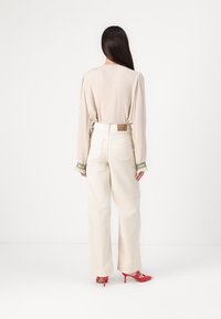 Camisa de manga larga de color beige claro con mangas fruncidas, combinada con pantalones vaqueros de pierna ancha en color crema que presentan una etiqueta VMD en la banda de la cintura y unos zapatos de tacón rojos.