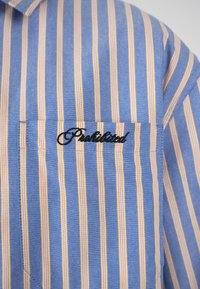 Chemise rayée verticale bleu et beige avec une poche poitrine brodée du mot « Prohibited » en lettres cursives noires.