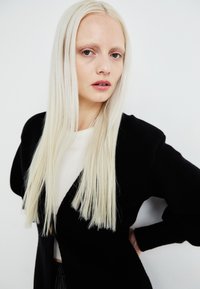 Black textured jacket over a white top, featuring long straight blonde hair and a neutral background. No notable patterns or accents.