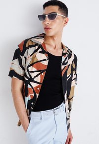Chemise à manches courtes avec un motif abstrait multicolore. Débardeur noir en dessous, associé à un pantalon bleu clair et une ceinture tressée.