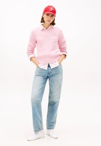 Rosa gestrickter Pullover mit strukturiertem Muster, weißes Hemd mit Kragen darunter, hellblaue Jeans und eine rote Mütze. Weiße Turnschuhe vervollständigen das Outfit.