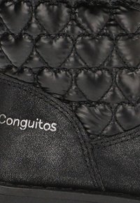 Tela negra acolchada con patrones de corazones y el logo "Conguitos" en relieve en una sección de cuero liso del calzado.