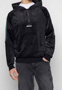 Felpa nera in velluto con mezza zip, strisce verdi ai lati e una patch logo sulla parte anteriore. Presenta una tasca a canguro e polsini a coste.