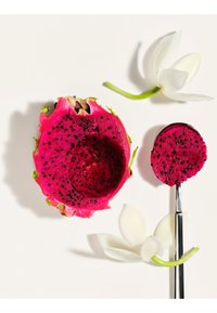 La moitié d’un fruit du dragon rose vif avec des graines noires, une cuillère tenant un morceau rond du fruit, et deux fleurs blanches sur un fond clair.