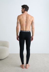 Cuecas longas pretas com bainha de marca, design ajustado, textura suave, usadas por uma figura masculina com tatuagens nas costas.