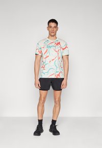 Colorful short-sleeve t-shirt with blue, red, and light green abstract pattern on a cream background. Paired with black shorts and shoes.