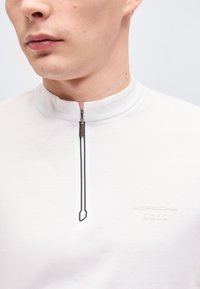 Κοντινό πλάνο ενός ατόμου που φοράει λευκό πουκάμισο με φερμουάρ, με κοντό γιακά και ανάγλυφο λογότυπο «Porsche BOSS» στο στήθος.