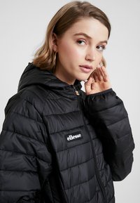 Doudoune noire avec un design matelassé, capuche attachée et fermeture éclair à l'avant. Présente un logo ellesse sur le côté gauche de la poitrine.
