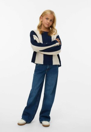 Pull en maille rayé bleu marine et crème avec une texture côtelée, associé à un jean en denim bleu à jambes larges. Les chaussures présentent des accents couleur crème.