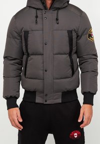 Gray padded jacket with a hood, featuring a front button closure, side pockets, and black accent details. Includes logo patch on sleeve.