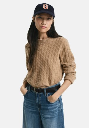 Beige Pullover aus Kabelstrick mit Rundhalsausschnitt und umgeschlagenen Ärmeln, kombiniert mit hoch sitzenden blauen Jeans und einem schwarzen Gürtel. Marinefarbene Mütze mit einem roten Buchstaben G.