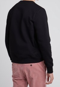 Person som bär en svart långärmad sweatshirt och rosa kordflöjlsbyxor, visad bakifrån mot en enfärgad ljus bakgrund.