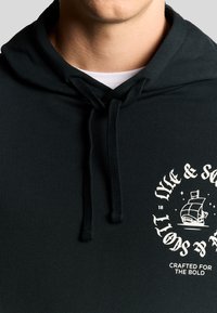 Felpa verde scuro con tasca frontale, caratterizzata da grafiche bianche a tema nautico e coulisse. Materiale in misto cotone con una texture morbida.