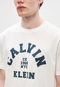 Valge crewneck t-särk, millel on tumesinise värviga graafiline print, mis kuvab "CALVIN KLEIN CK 1968 NYC". Kangas näeb välja pehme ja sile ning sellel on lühikesed varrukad.
