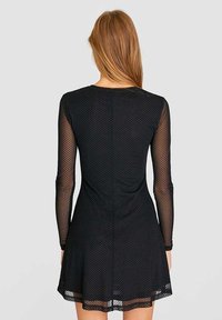 Vestido negro con mangas largas y transparentes de lunares, con un corsé ajustado que se abre en un dobladillo acampanado. Tela de malla texturizada con un forro suave.