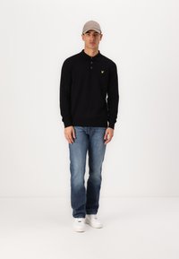 Polo negru cu mâneci lungi și un logo galben, asortat cu blugi albaștri decolorati și adidași albi. Modelul poartă o șapcă gri deschis.