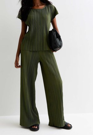 Frau trägt olivgrünes, plissiertes Kurzarmshirt und weite Hosen, hält eine schwarze Handtasche, mit schwarzen Sandalen.