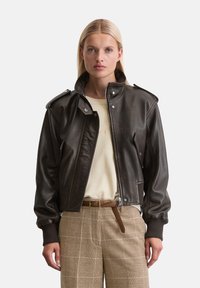 Braune Lederjacke mit Reißverschluss, Kragen und Schulterklappen. Kombiniert mit einem beigen Oberteil und braunen karierten Hosen.