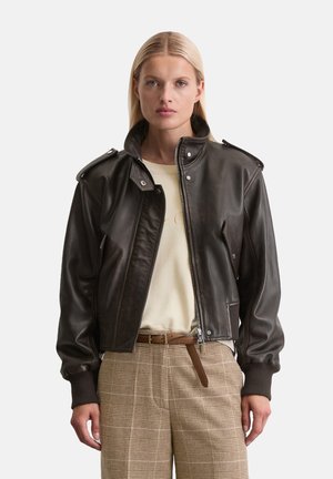 Braune Lederjacke mit Reißverschluss, Kragen und Schulterklappen. Kombiniert mit einem beigen Oberteil und braunen karierten Hosen.