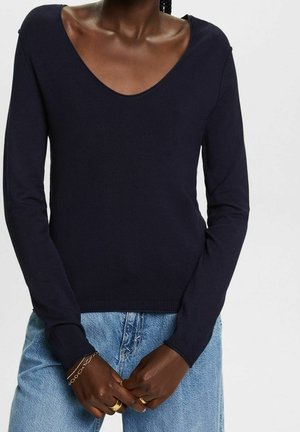 Osoba nosząca ciemnogranatowy sweter z dekoltem w serek i jasnoniebieskie dżinsy, ręce złożone, nosi złote pierścionki i bransoletkę z łańcuszkiem.