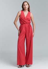 Rode, mouwloze jumpsuit met een wikkeldesign, cinch taille en brede pijpen. Gemaakt van een zachte stof met een minimale textuur. Zwarte sandaaltjes met hoge hakken.