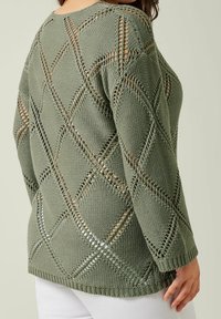 Kobieta w luźnym oliwkowym swetrze dzianym z diamentowym wzorem z otworami oraz białych spodniach, widoczna od tyłu.