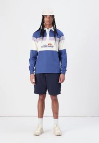 Sininen ja valkoinen pitkähihainen polo, jossa on raidoitus, varustettuna "ellesse"-logolla. Yhdistettynä tummansinisiin shortseihin ja beigeihin tennareihin.