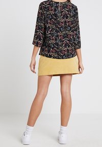 Haut noir à motif floral avec des manches 3/4, doté d'une fermeture éclair au dos ; associé à une jupe en velours côtelé jaune. Baskets blanches et chaussettes.