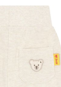 Helle beige, strukturierte Stoffhose mit einer Gesäßtasche, auf der ein gesticktes Bärengesicht zu sehen ist, sowie einem gelben Etikett an der Seite.