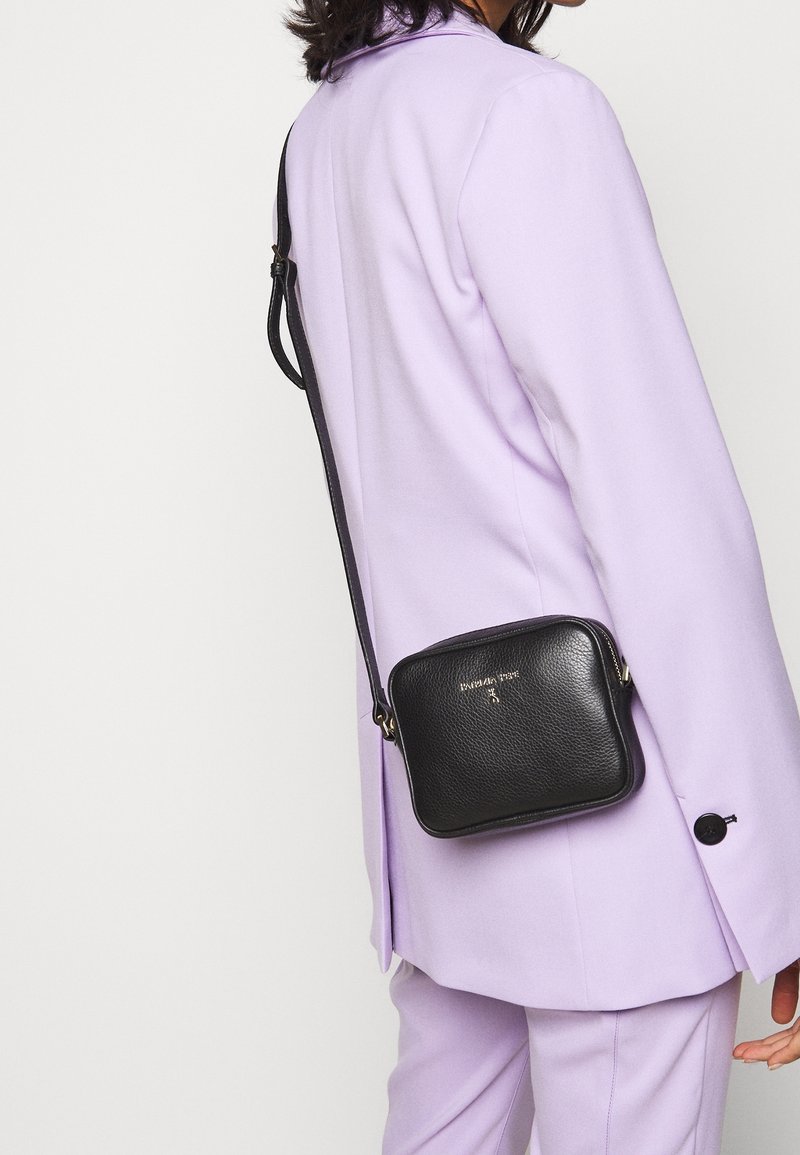 Černá kožená crossbody taška čtvercového tvaru, se zipem a dlouhým popruhem. Na přední straně má jemně embosované logo.