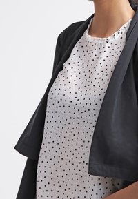 Veste noire ample portée sur un chemisier blanc à pois noirs, avec un col froncé et un tissu léger.