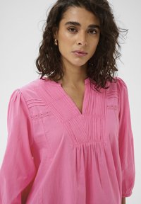 Blouse rose à col en V, avec des plis verticaux et des détails en dentelle sur les épaules. Manches longues légèrement bouffantes offrant une coupe décontractée.