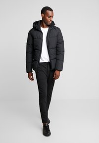 Black puffer jacket with a hood, featuring a quilted design. Paired with black pants and black sneakers. Model standing against a neutral background.