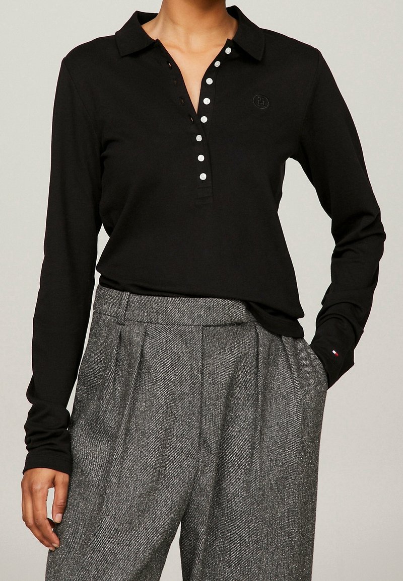 Langärmeliges schwarzes Poloshirt mit Kragen, silbernen Knöpfen und einem dezenten Logo, kombiniert mit grauen, strukturierten, hoch taillierten Hosen.