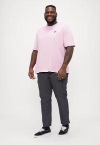 Tricou din bumbac roz deschis, croială lejeră, mâneci scurte, logo galben pe piept; asortat cu pantaloni cargo gri închis cu buzunare laterale și adidași negri.