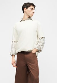 Νέος άνδρας που φοράει στρωμένο μπεζ καρό πουκάμισο κάτω από ένα εκρού κοντομάνικο μπλουζάκι και καφέ παντελόνια με φαρδιά μπατζάκια, κοιτάζοντας προς τα πλάγια.