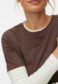 Καστανό βαμβακερό t-shirt με στρογγυλή λαιμόκοψη και κοντά μανίκια, φορεμένο πάνω από ένα λευκό μακρυμάνικο πουκάμισο, που αναδεικνύει απαλό ύφασμα.