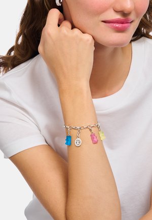 Silberkette mit drei bunten Gummibärchen-Anhängern (blau, rosa, gelb) und einem kleinen ovalen Anhänger, getragen am nackten Handgelenk.